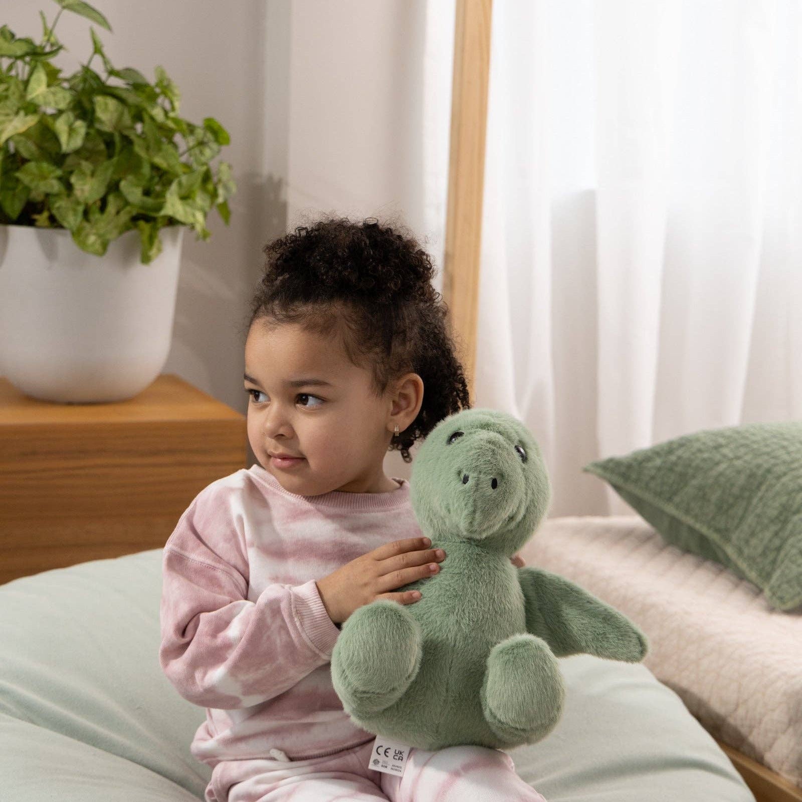Child sitting on a cushion holding a green teddy bear in a room with a plant and bed in the background.