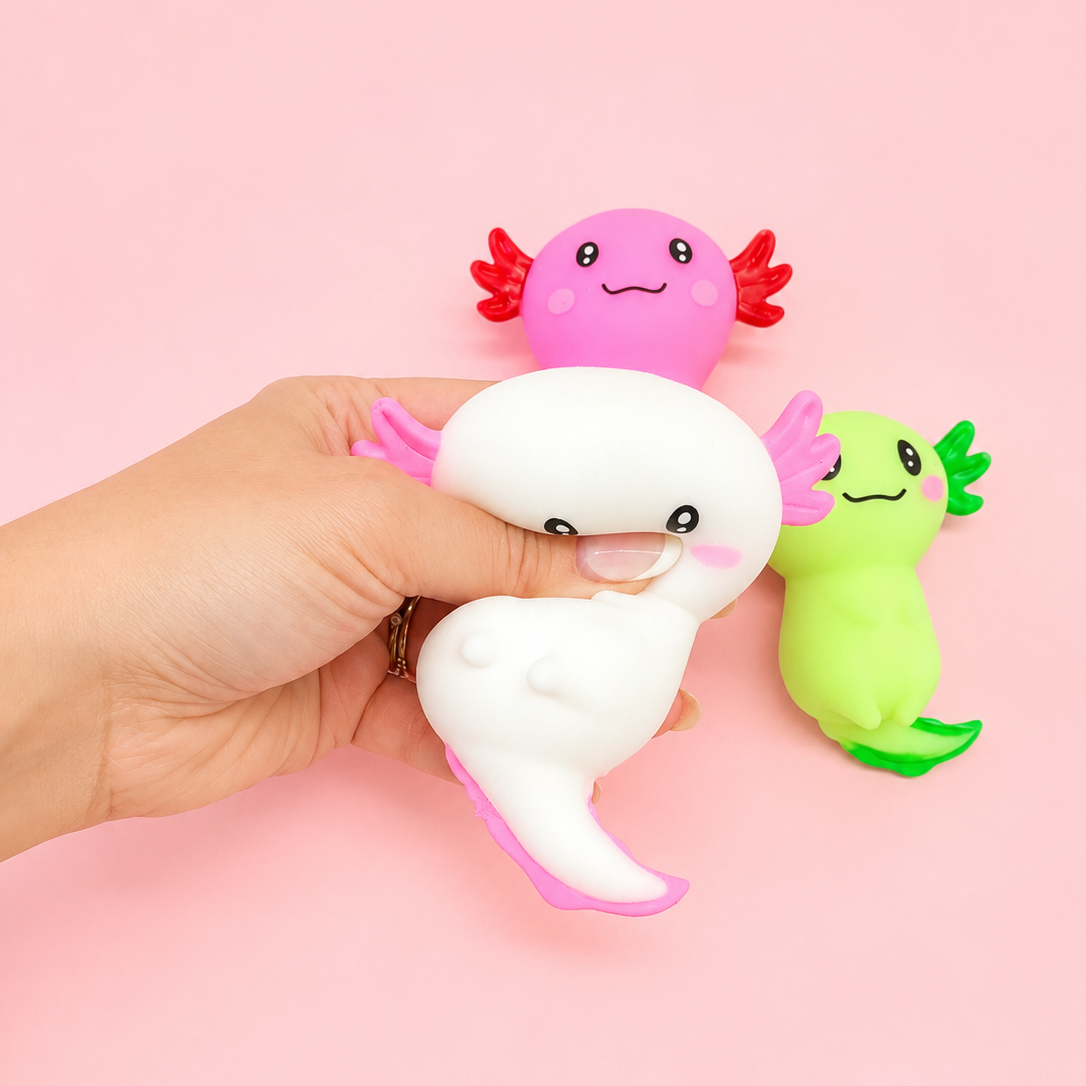 A hand holds a white and pink axolotl-shaped squishy toy, with two smaller axolotl toys in pink and green standing behind it against a solid pink background.