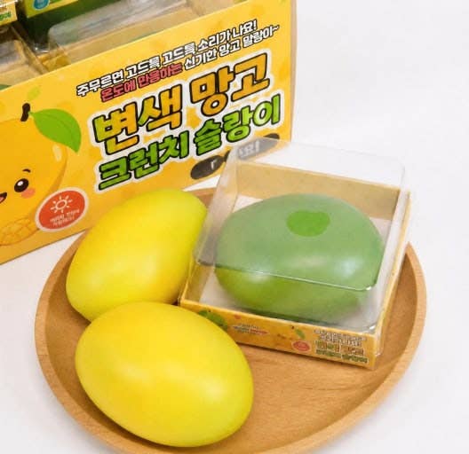 A yellow mango-shaped novelty item displayed on a wooden plate with its packaging box in the background. The box is yellow with green and black Korean text and a cartoon mango illustration.