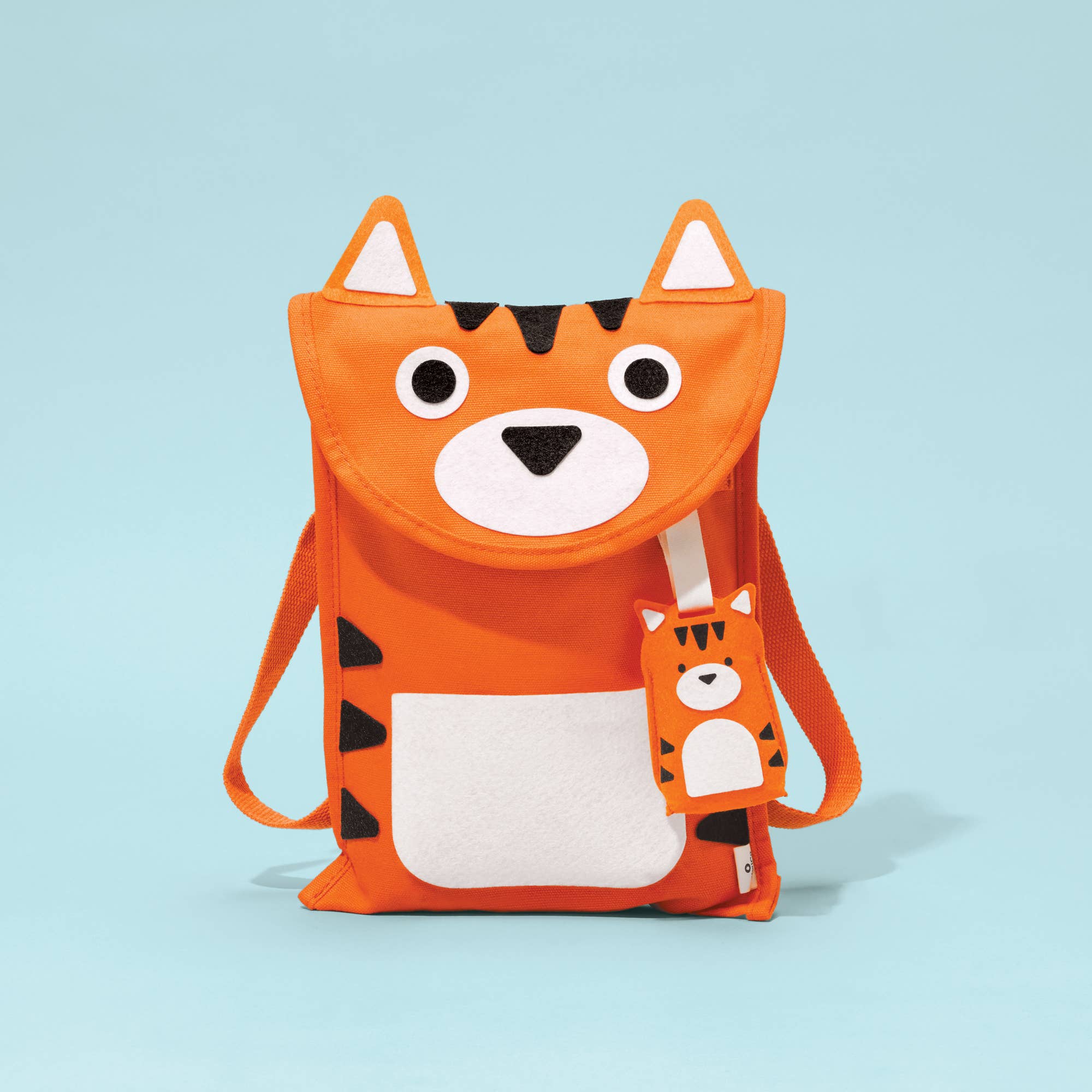 An orange cat-shaped backpack with a white front pocket, black stripes, and a matching cat-shaped luggage tag, set against a light blue background.