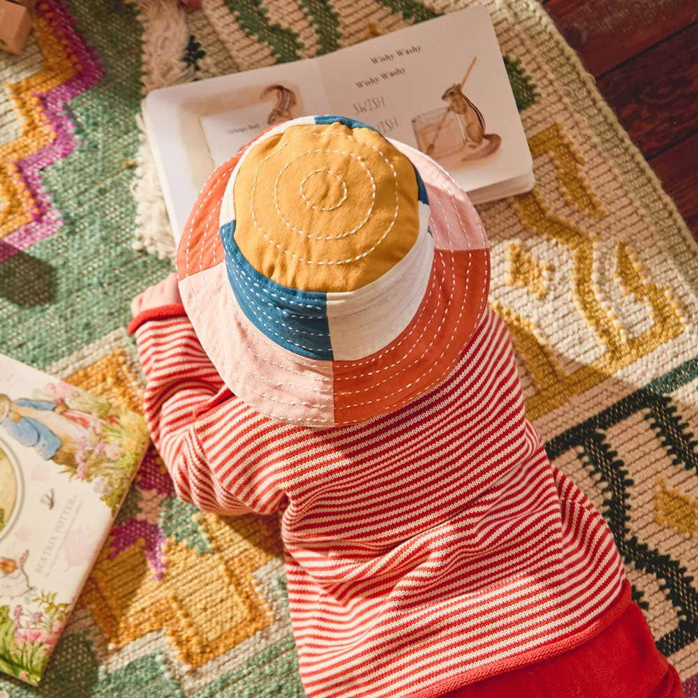 Child wearing a colorful patchwork hat and red outfit on a patterned rug with books around