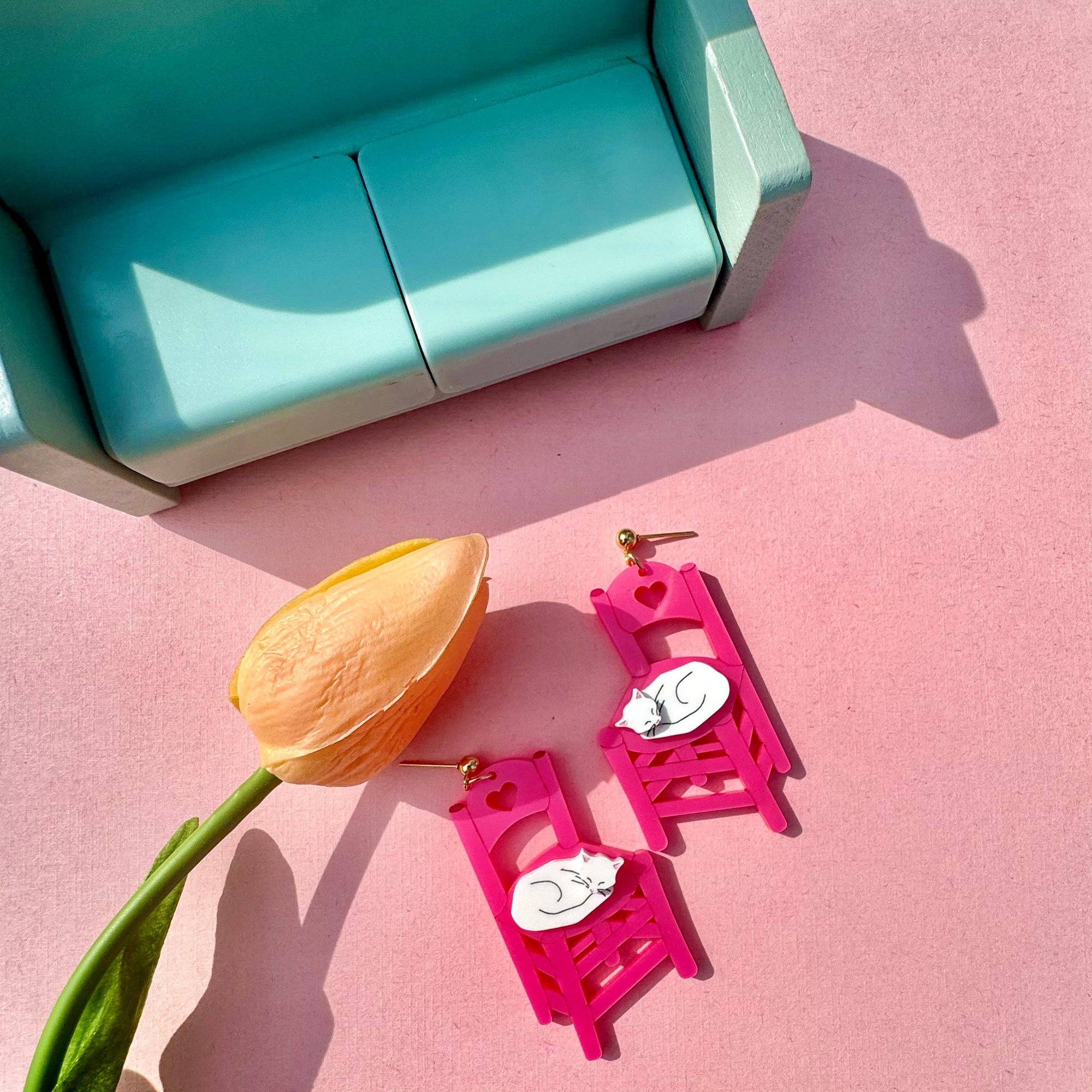 Pink earrings with a cat design on a pink surface next to a tulip and turquoise box.