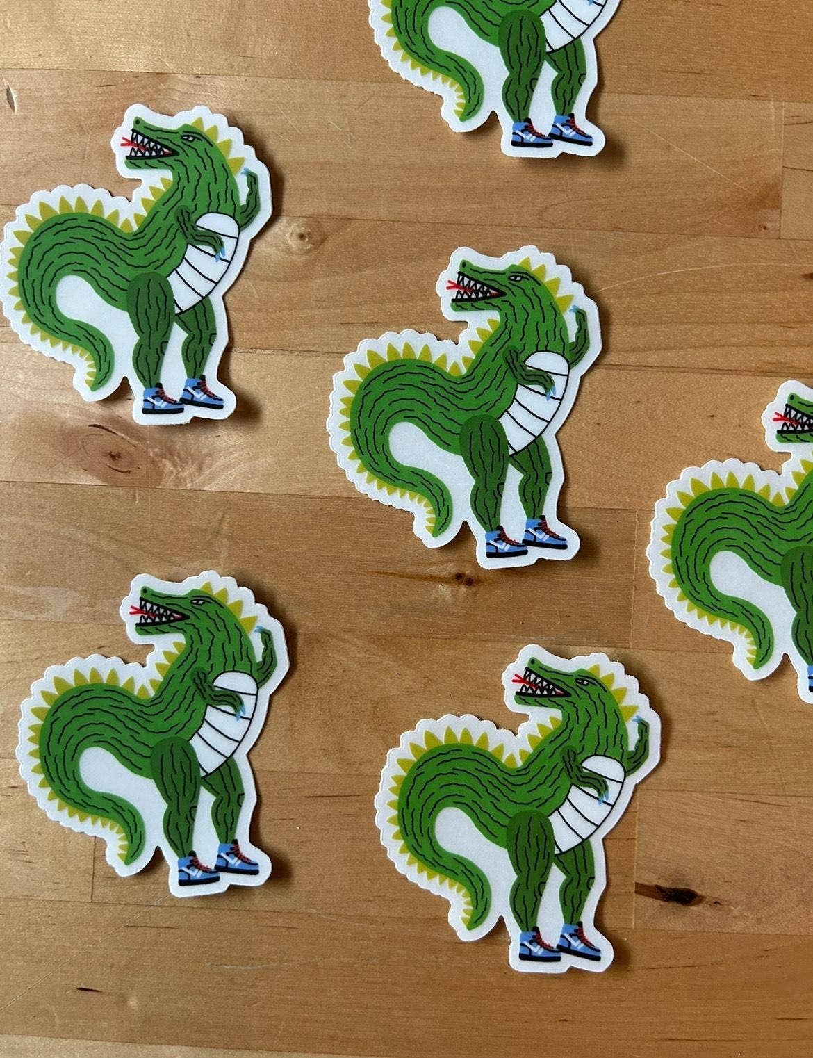 Several green cartoon dinosaur stickers with white borders are arranged on a light wood surface.
