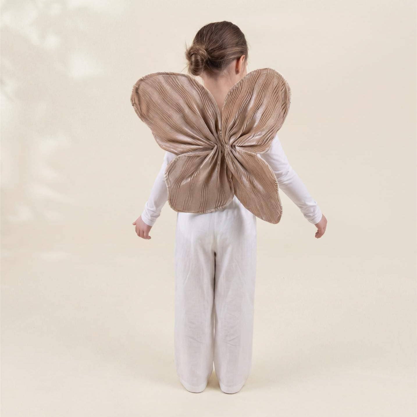 A child standing with their back to the camera, wearing a white long-sleeve top and white pants, with a large brown butterfly wing costume attached to their back, against a warm beige background.