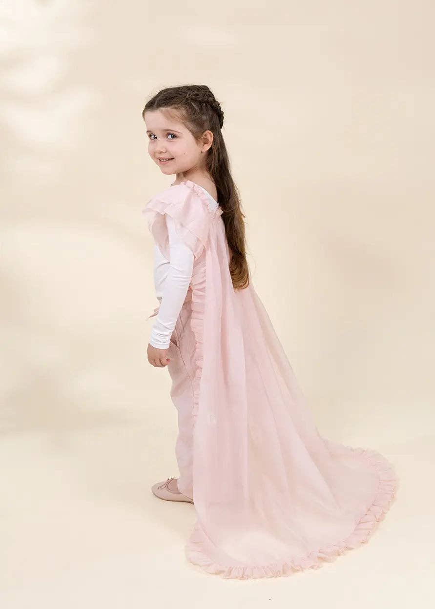 A young girl with long brown hair in a half-up style, wearing a light pink ruffled cape dress over a white long-sleeve top, standing against a warm beige studio background.