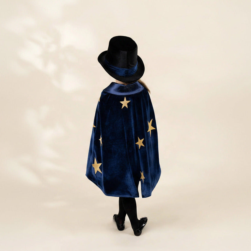 A child shown from the back wearing a dark blue velvet cape with gold stars and a matching black top hat, standing against a plain warm beige studio background.