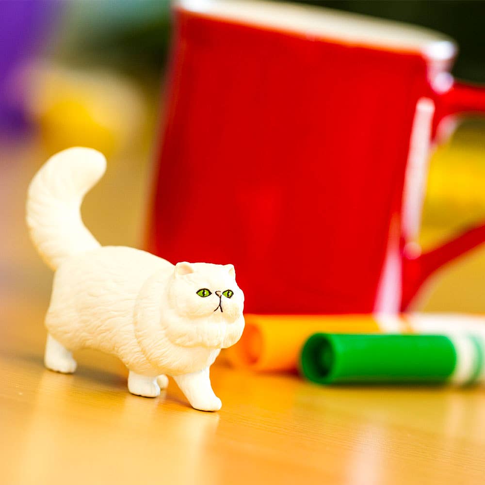 White cat figurine on a wooden surface with a red mug and colorful objects in the background.