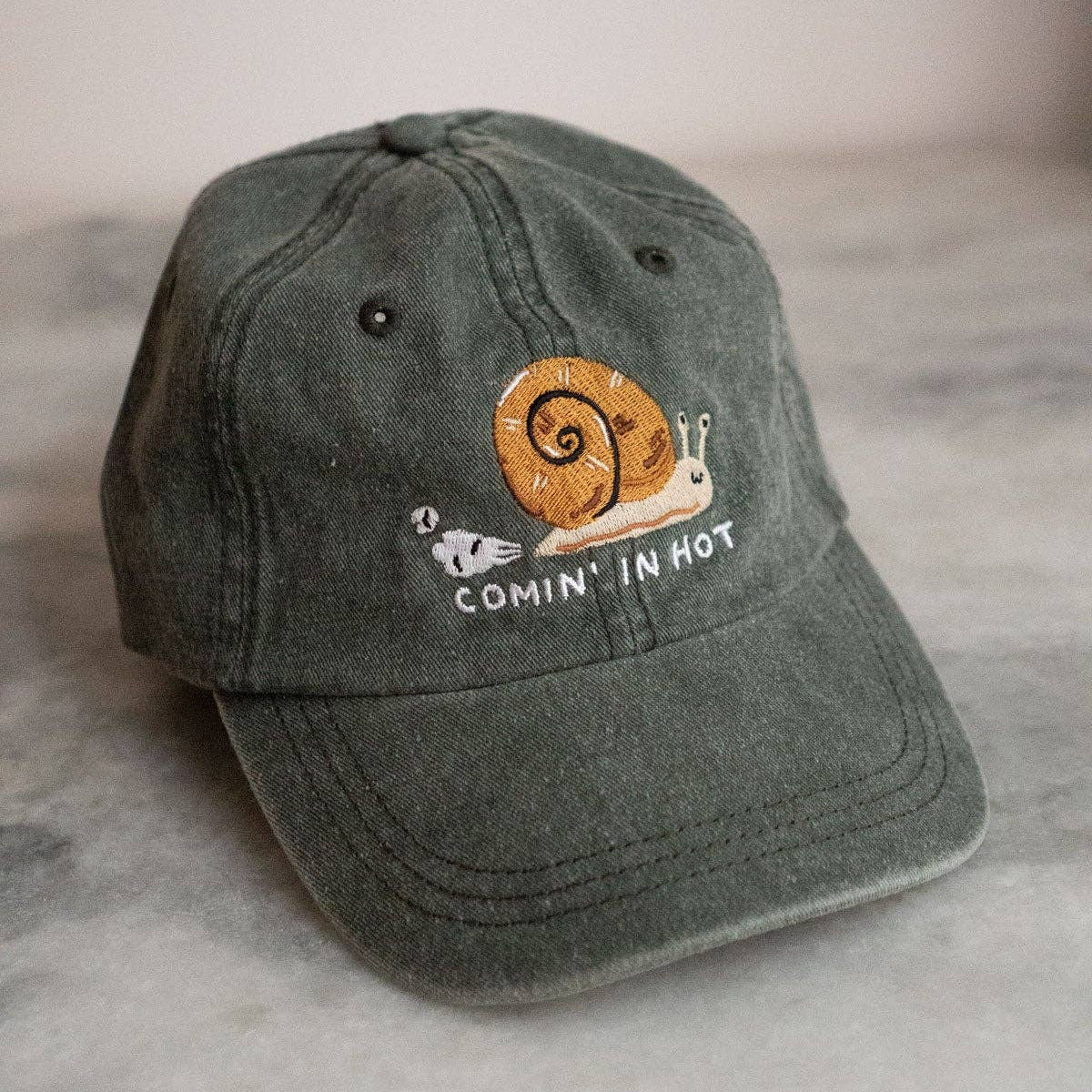 Green cap with a snail design and 'Comin' in Hot' text on a marble surface.