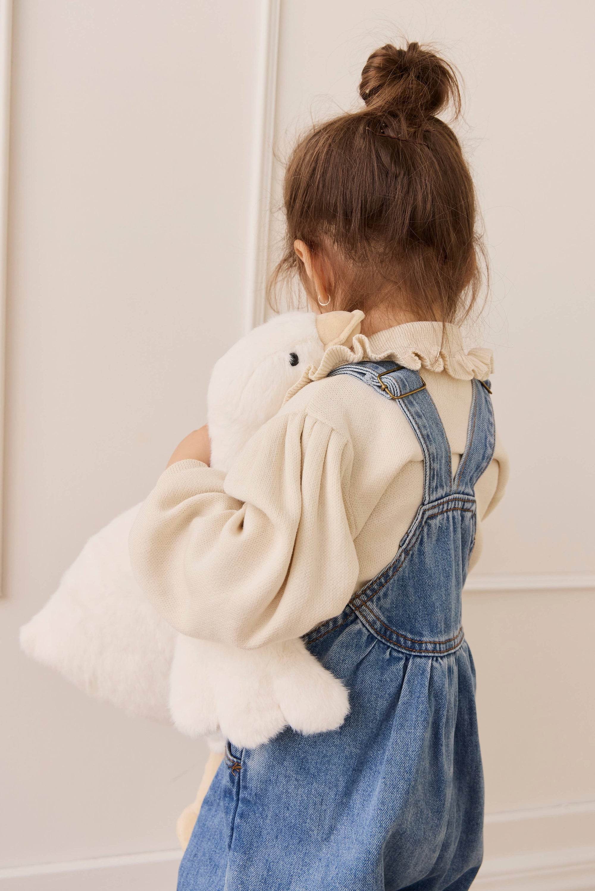 Child wearing a denim dress holding a white teddy bear against a plain background