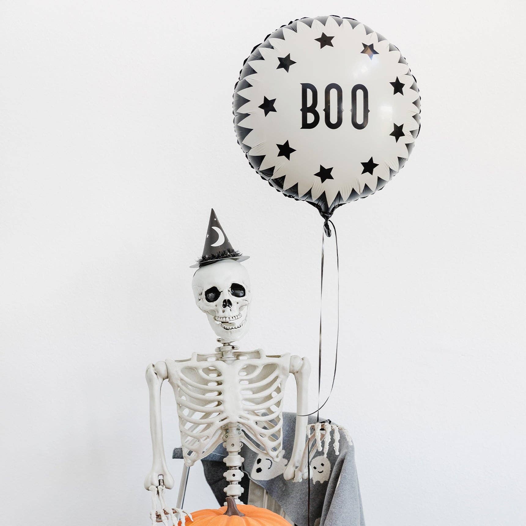 Skeleton holding a pumpkin with a 'BOO' balloon on a white background