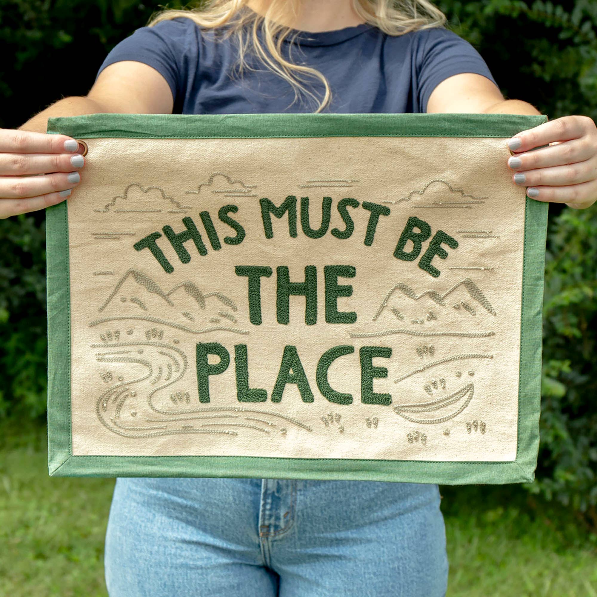 Person holding a wooden sign with 'This Must Be the Place' text and nature illustrations.