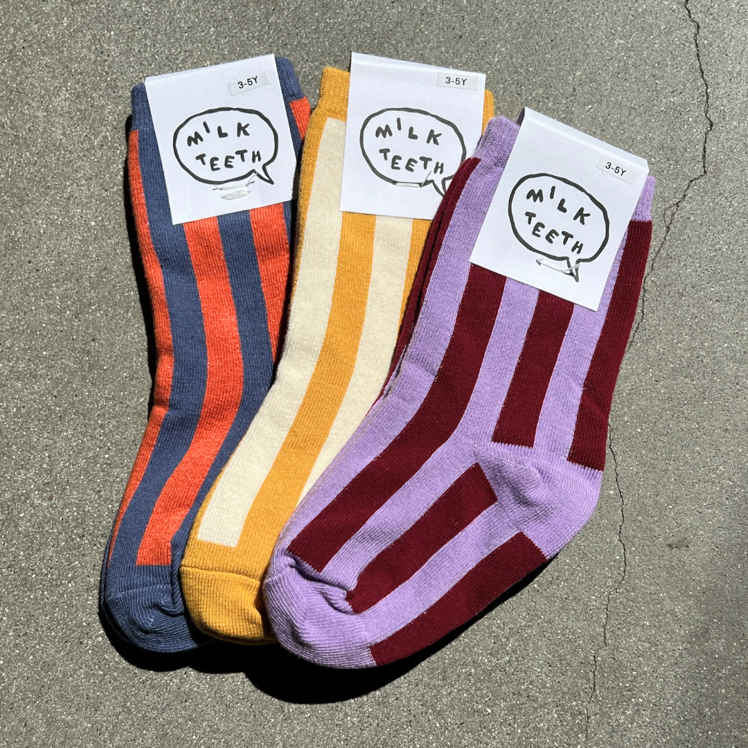 Three pairs of striped socks with 'Milk Teeth' labels on a concrete surface.