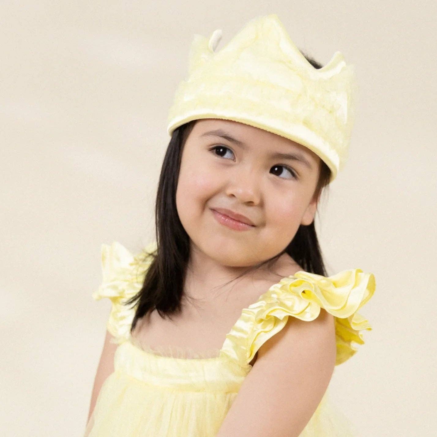 A young girl wearing a light yellow princess costume dress with ruffled shoulder straps and a matching yellow crown, photographed against a plain light beige background.