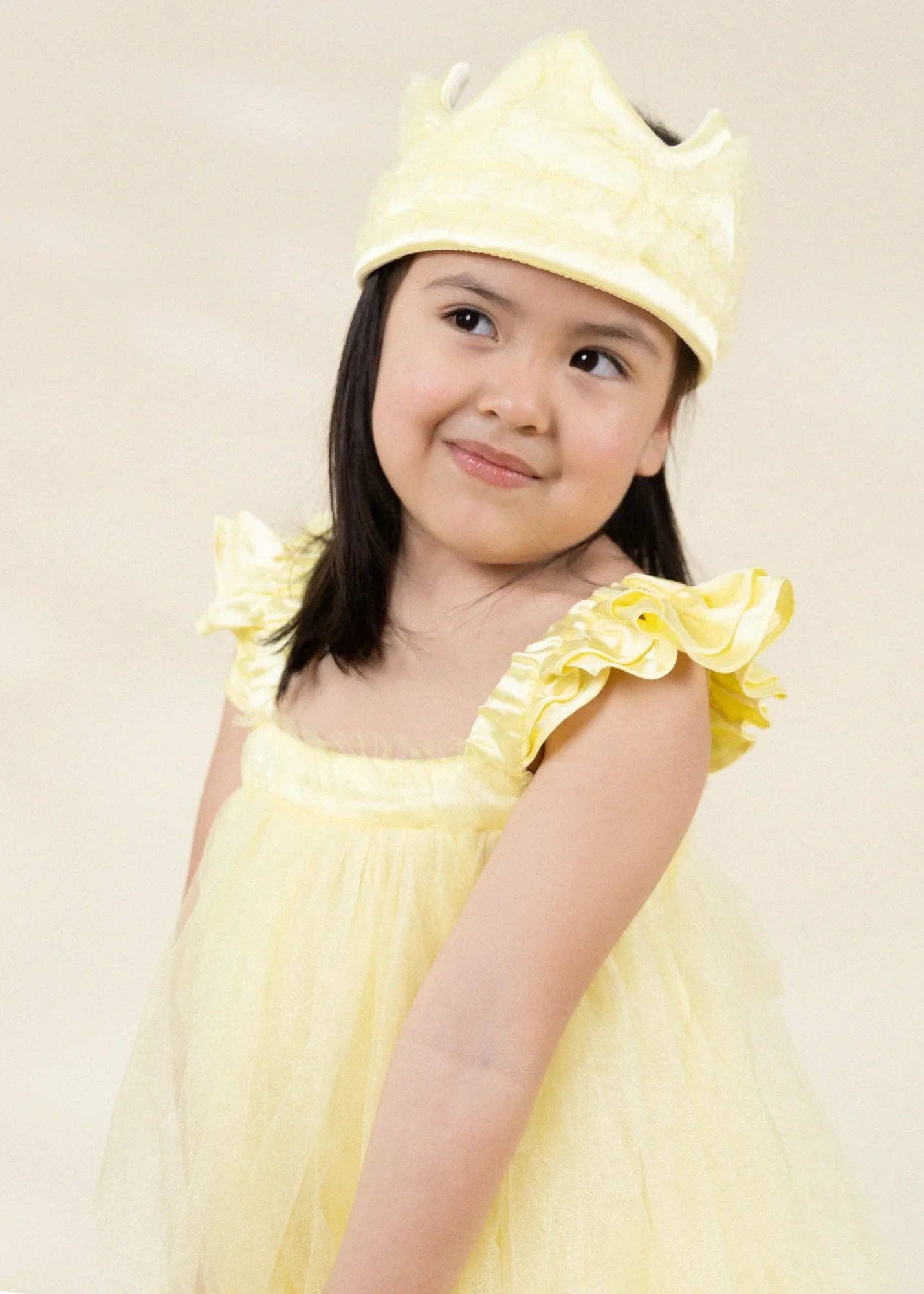 A young girl wearing a light yellow princess costume dress with ruffled shoulder straps and a matching yellow crown, photographed against a plain light beige background.