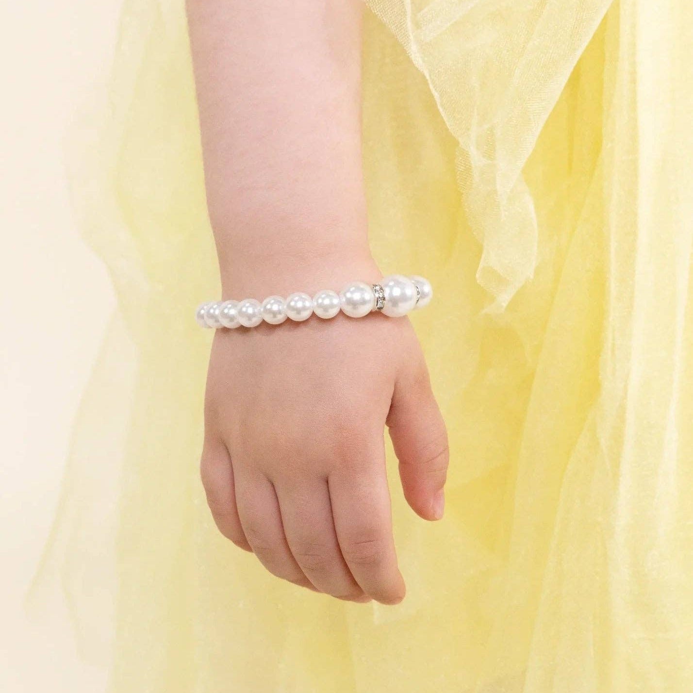 A close-up of a child's hand and wrist wearing a beaded bracelet made of white faux pearls, styled with a soft yellow tulle dress against a light cream background.