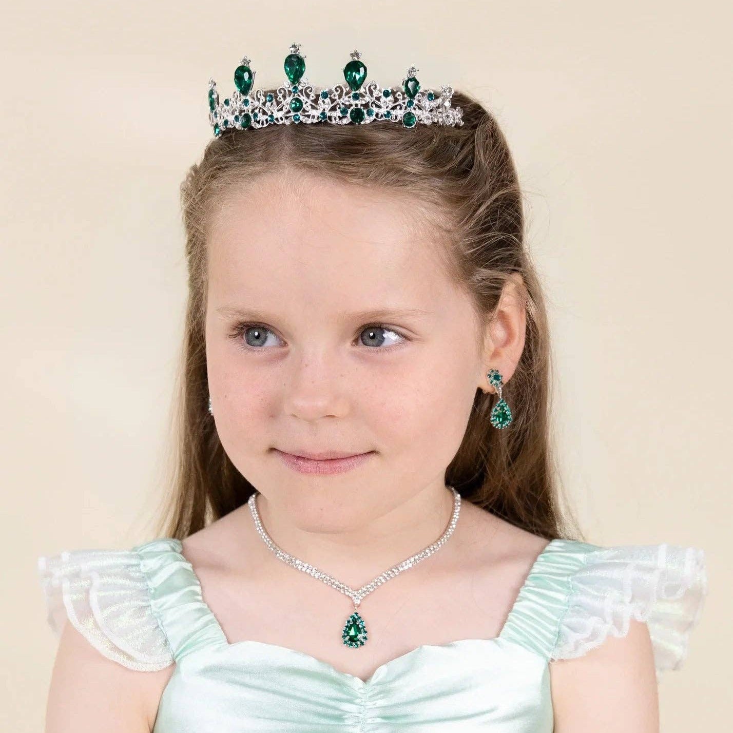 A young girl with brown hair wearing a silver tiara with green gemstones, a matching necklace, and earrings, paired with a mint green satin dress, set against a solid beige background.