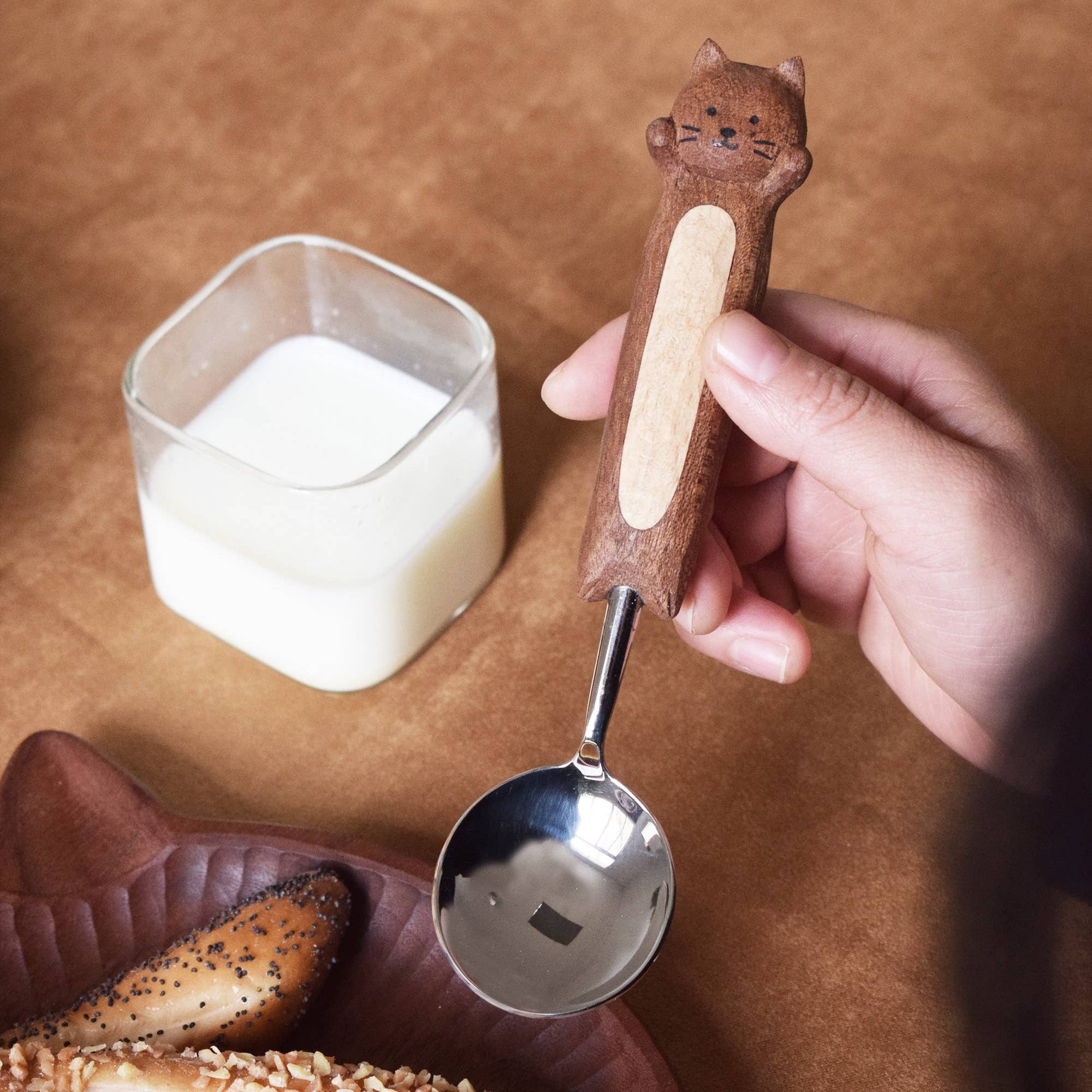 Hand holding a cat-shaped wooden spoon over a container of sugar on a wooden surface with pastries.