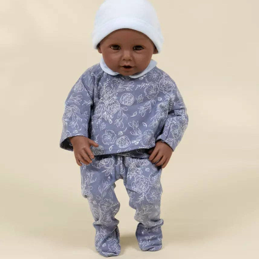 A baby doll wearing a light blue and white floral patterned two-piece outfit with a matching white bonnet, standing against a solid beige background.
