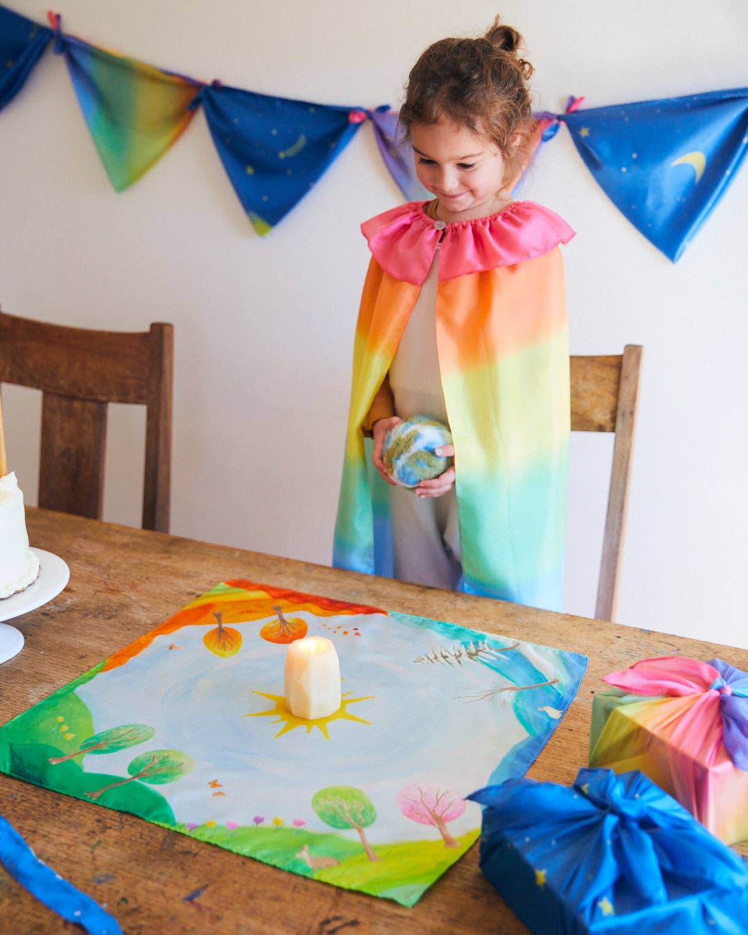 Child in a rainbow dress standing at a table with colorful decorations and a birthday cake.