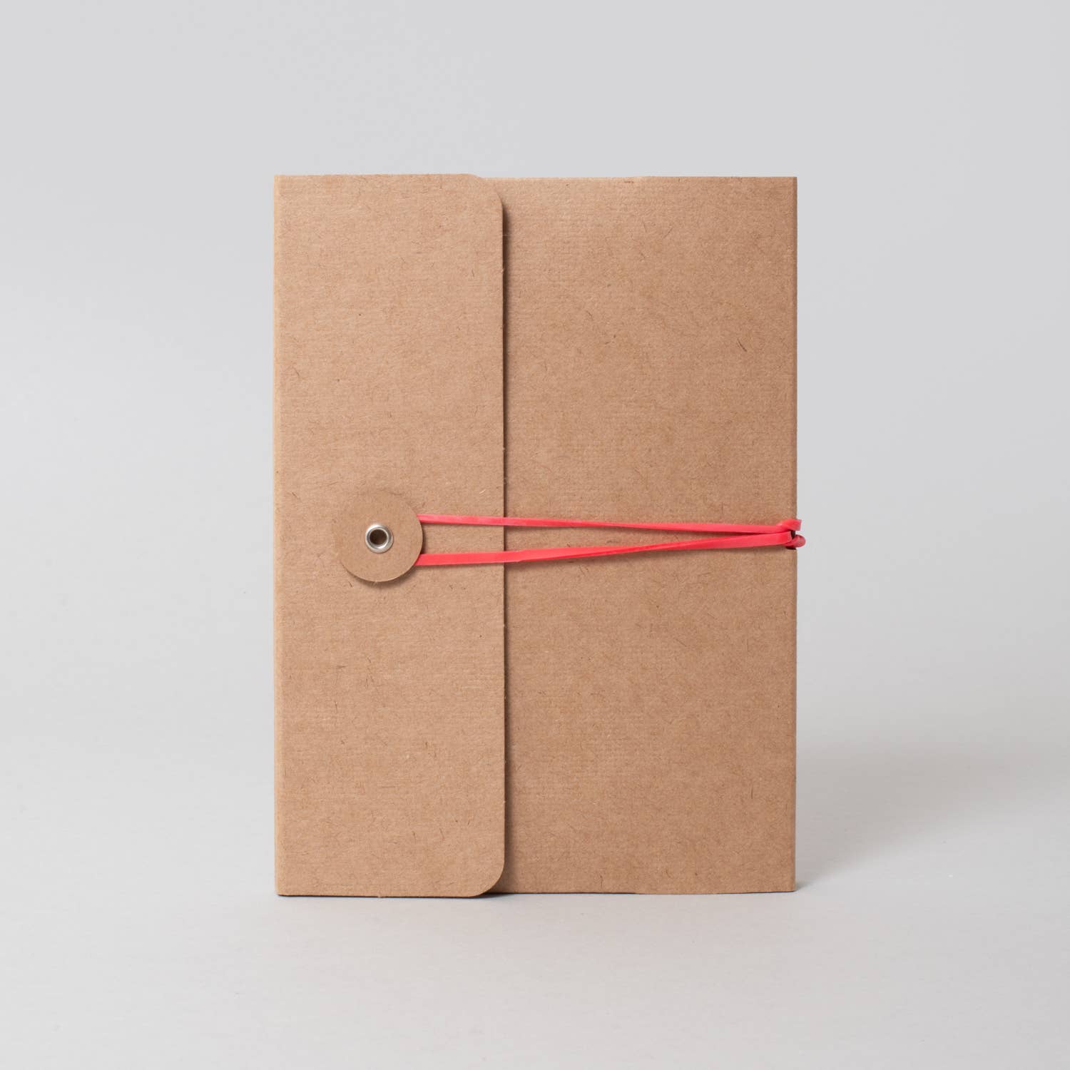 A kraft paper notebook or journal with a vertical flap closure, secured by a thin red elastic string and a small metal snap button, shown on a light gray background.