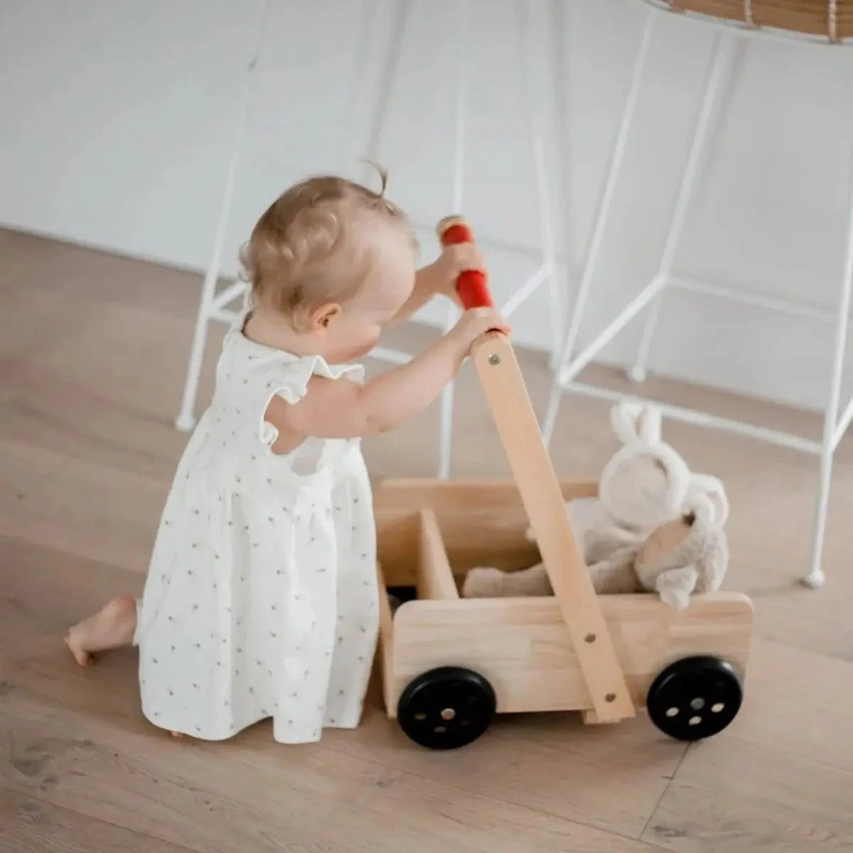 A baby in a white polka-dot dress pushes a natural wood toy wagon with black wheels and a red handle on a light wood floor. A white plush bunny sits inside the wagon.