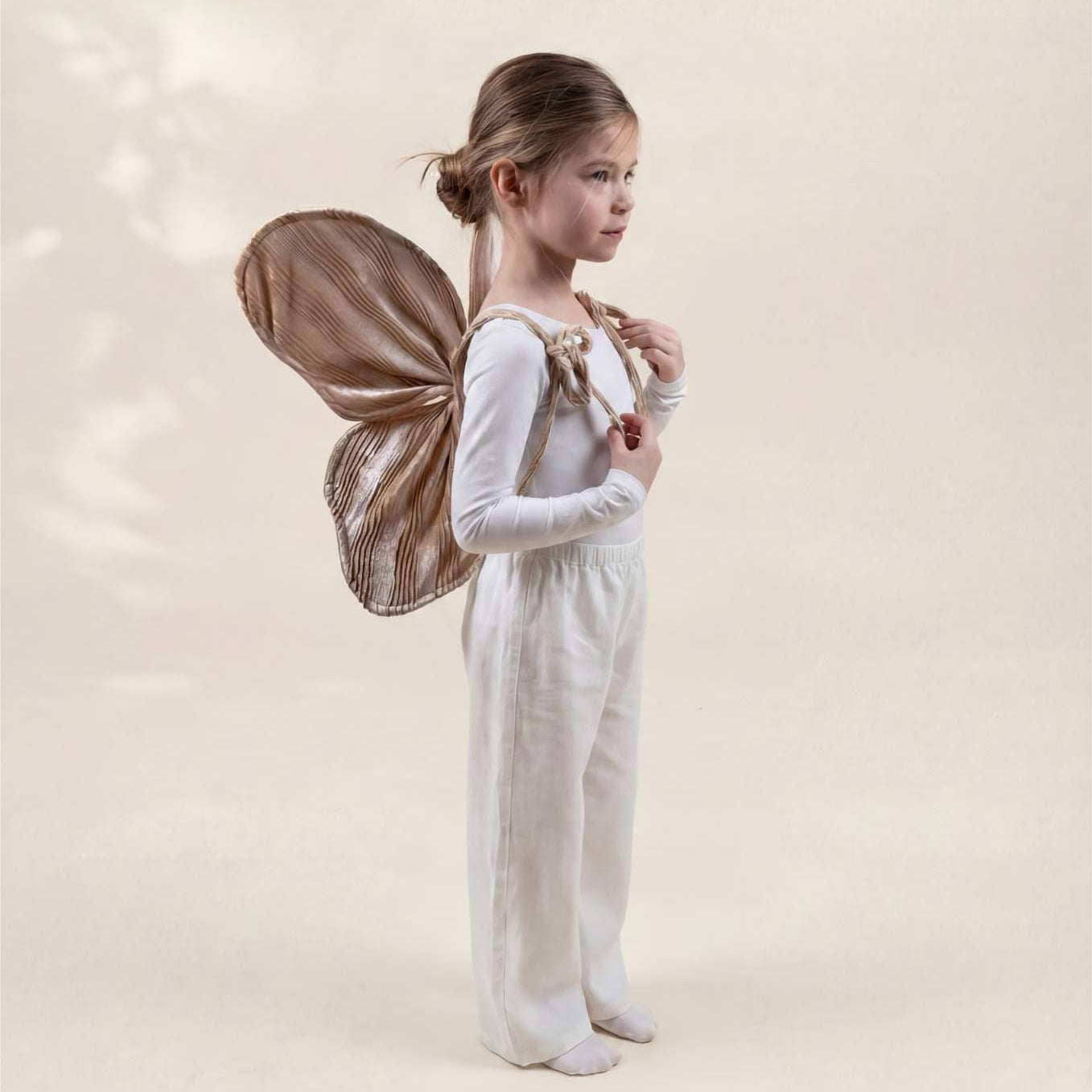 A young girl stands in profile wearing a white long-sleeve jumpsuit and barefoot, with large brown fairy wings attached to her back, against a soft warm beige studio background.