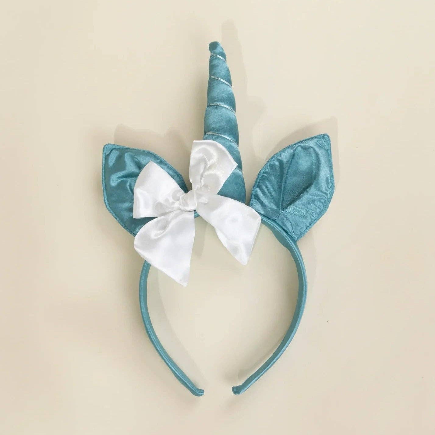A unicorn headband with a shiny teal blue horn, matching ears, and a white satin bow, displayed on a solid light beige background.