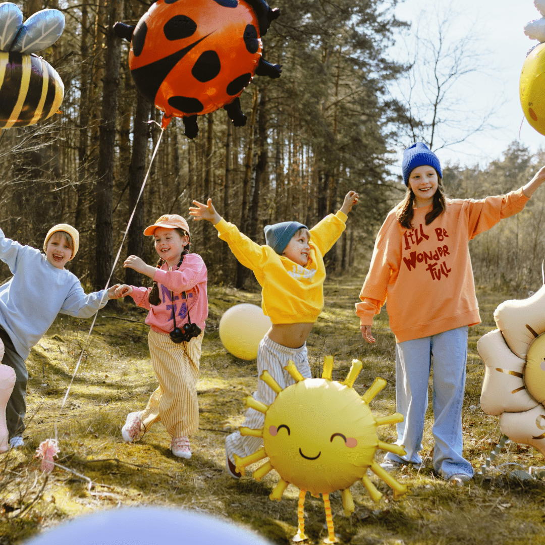 Four children play outdoors in a wooded area with colorful inflatable balloons, including a ladybug, sun, bee, and flower shapes. The children are wearing bright casual clothing and smiling, with trees and grass visible in the background.