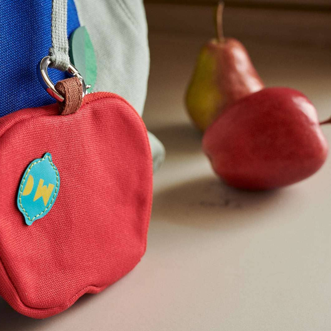 A red apple-shaped fabric pouch with a small blue and yellow patch, hanging from a blue bag next to two pears on a tabletop.