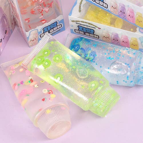 Three translucent squeeze tubes filled with glittery slime or decorative liquid, arranged on a pale pink surface. The tubes are pink, green, and blue, decorated with small cartoon and floral motifs.