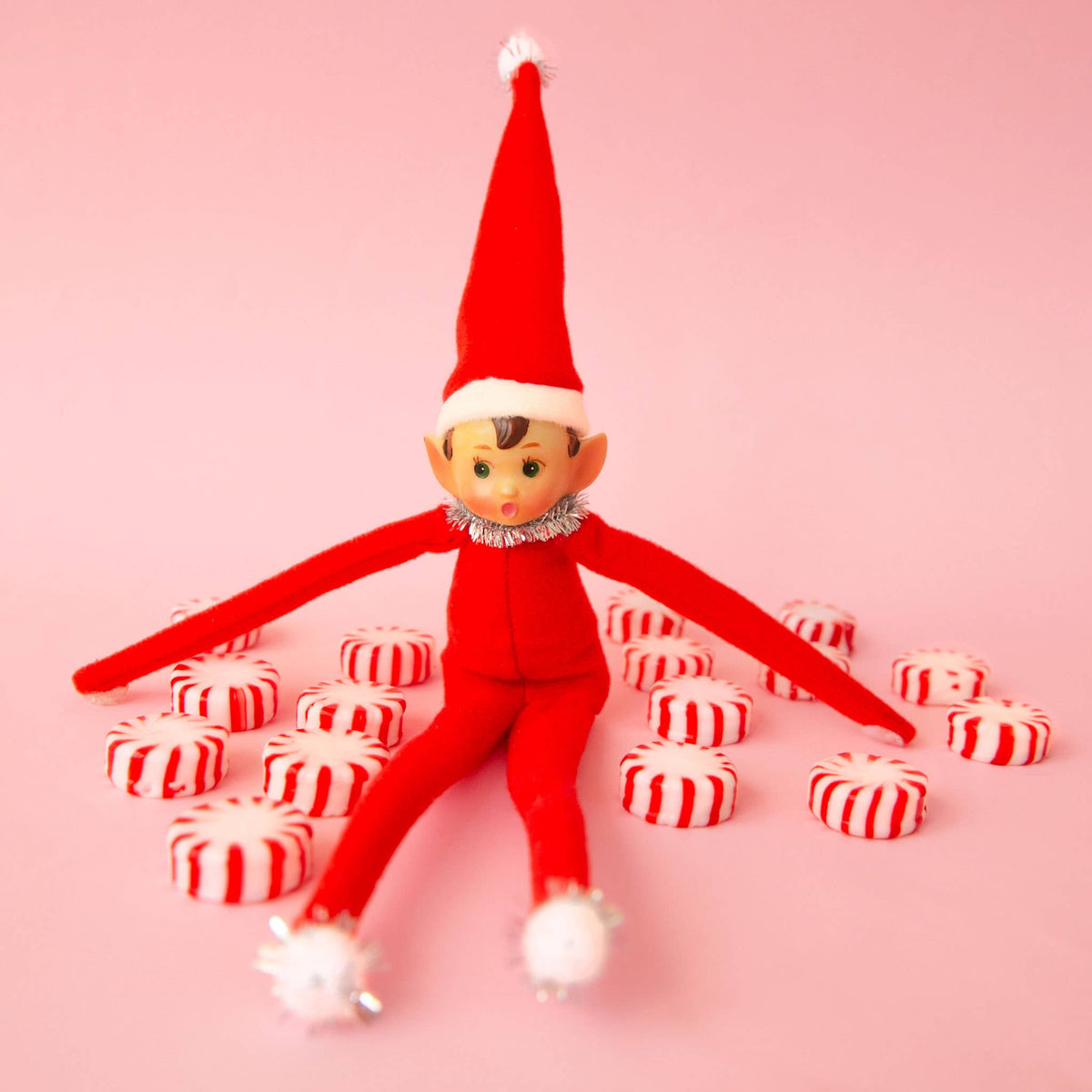 A red elf doll wearing a tall red hat with a white pom-pom and a silver collar, posed with red-and-white striped candy pieces on a solid pink background.