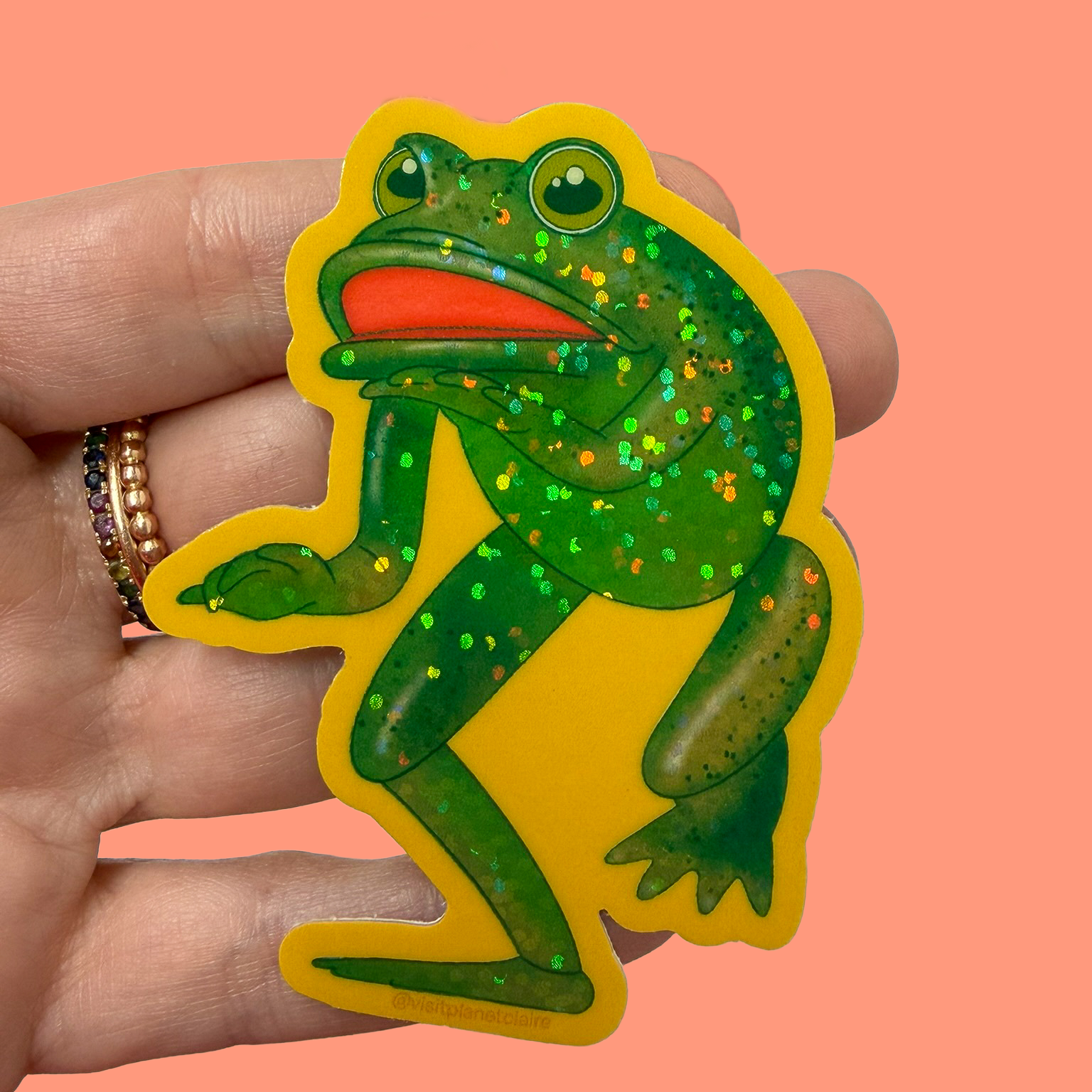 Hand holding a green frog sticker with a pink background