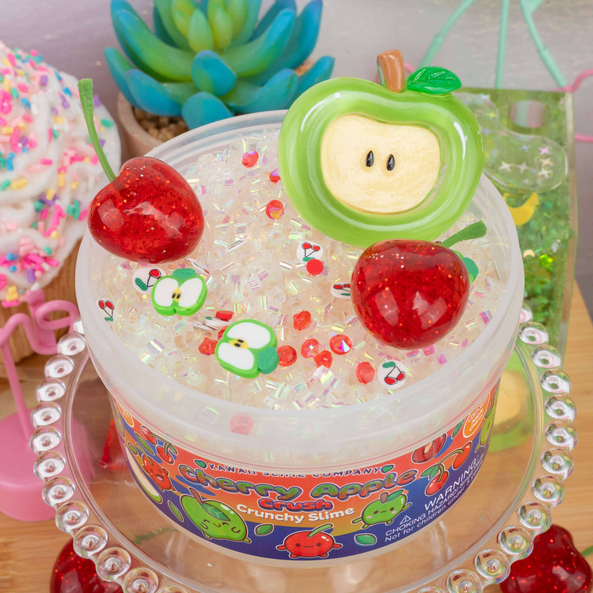 A tub of Crunchy Slime filled with clear glittery beads and topped with two red glittery cherries and green apple-shaped charms, displayed on a clear stand with colorful background props.