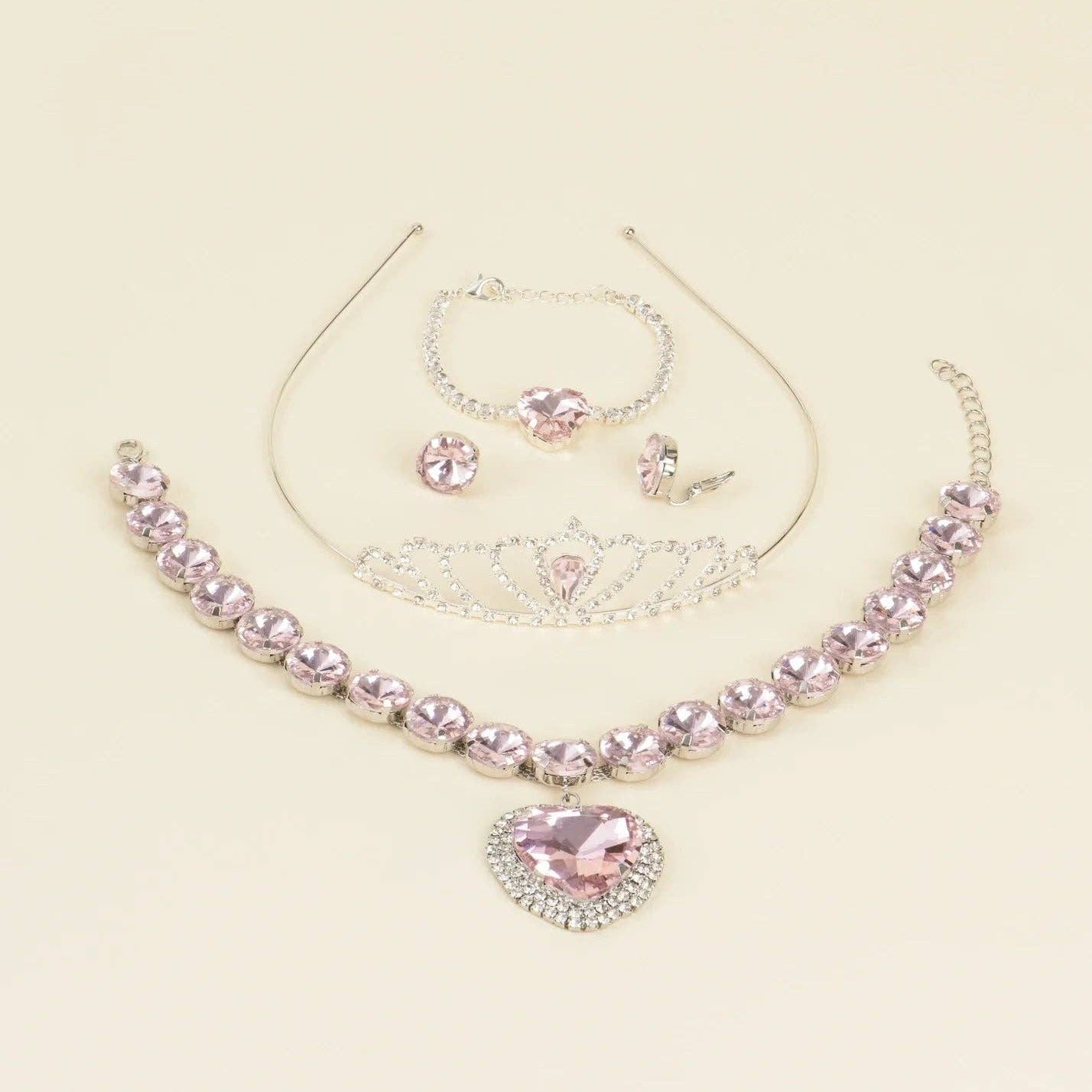 A pink and silver jewelry set on a light cream background, featuring a rhinestone-embellished collar necklace with a heart-shaped pendant, a tiara, a bracelet, and matching stud earrings.