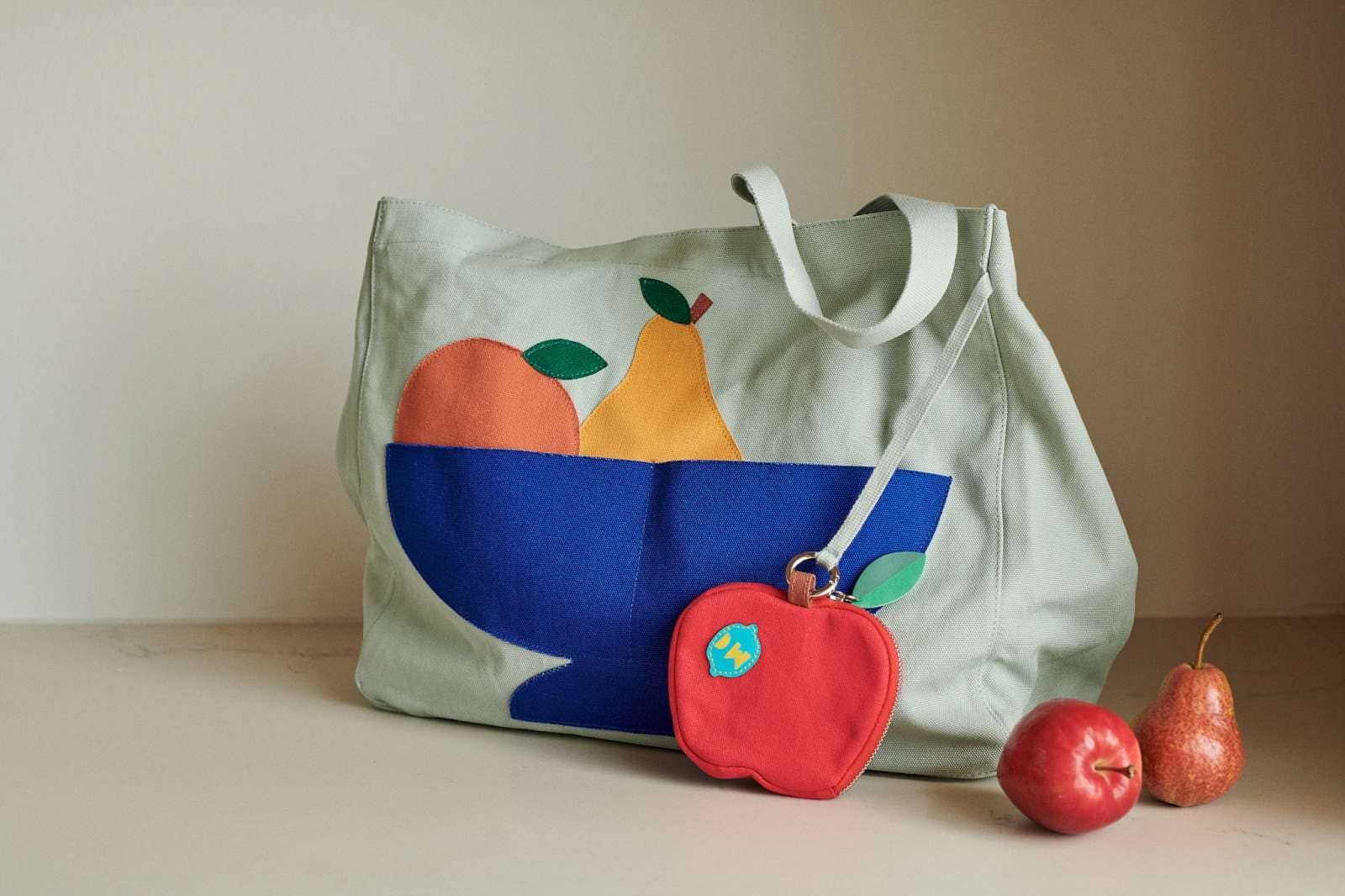 A canvas tote bag with a fruit-and-bowl applique design in sage green, blue, yellow, and orange, shown on a tabletop with two small fruit-shaped accessories and two whole fruits beside it.