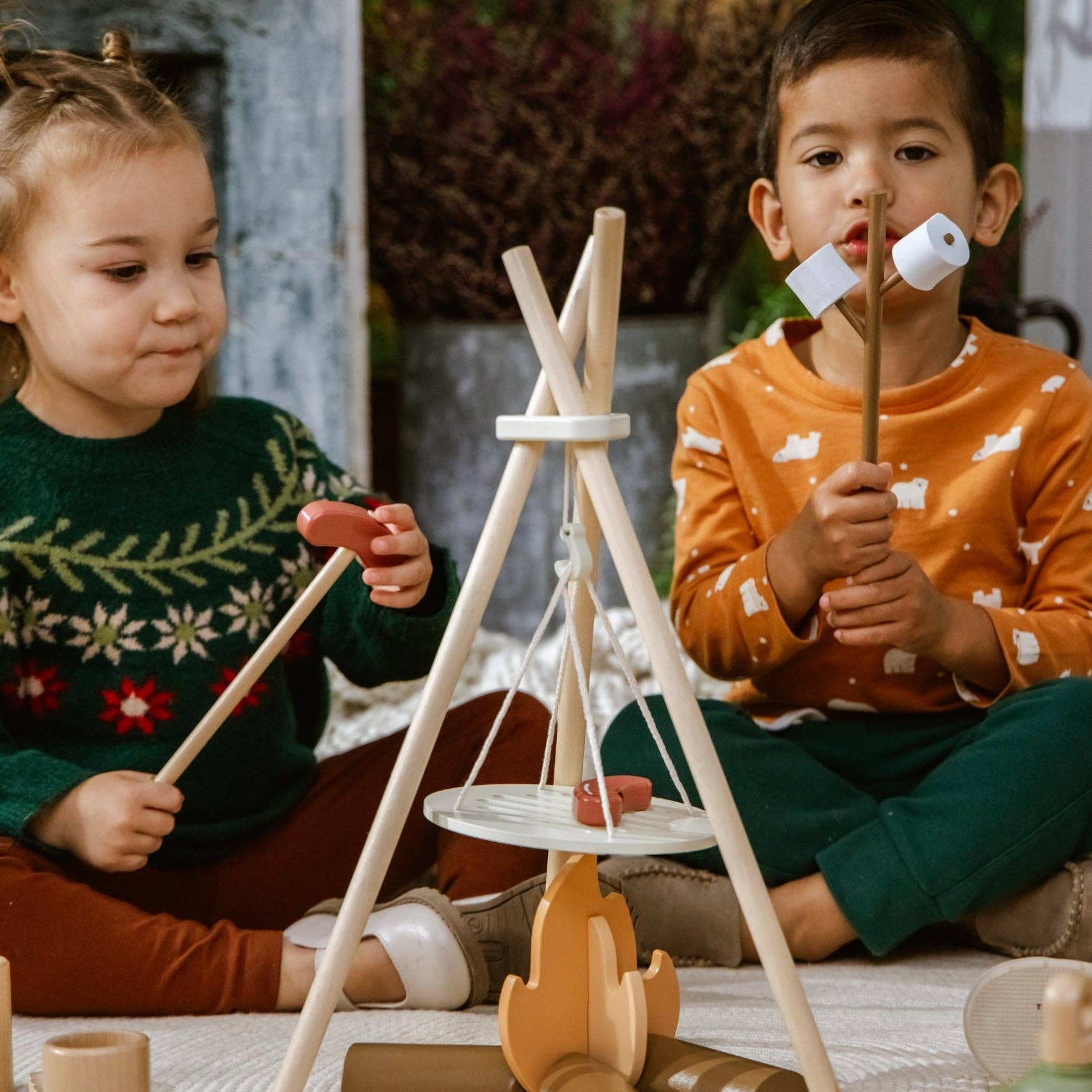 Two children playing with wooden toys including a teepee and campfire.