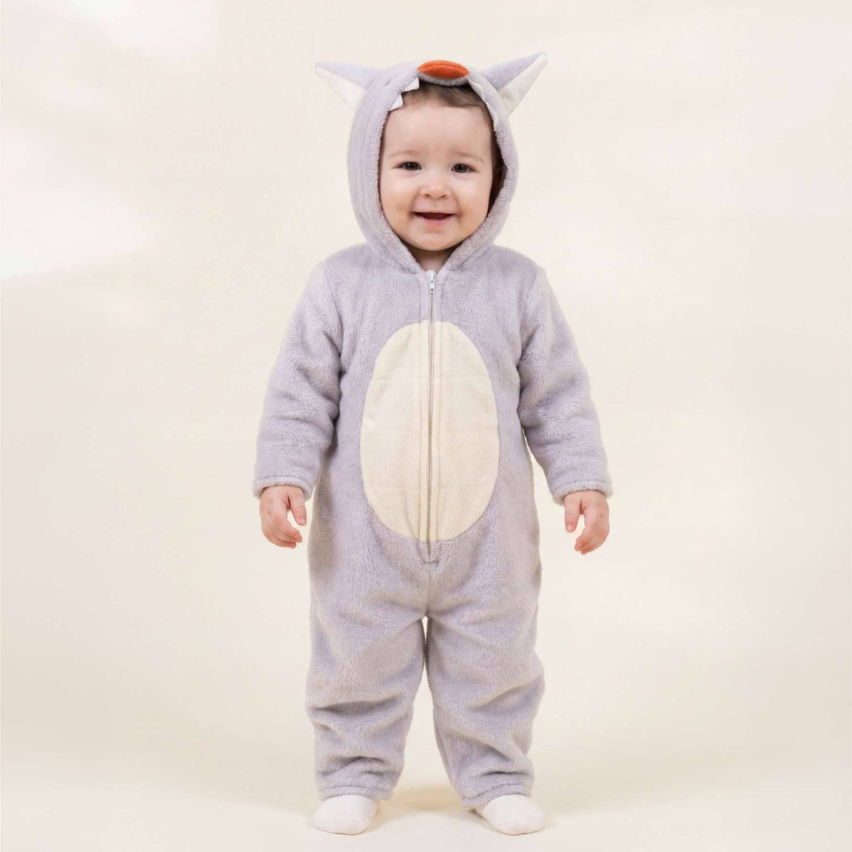 A baby wearing a light gray animal onesie with a white belly patch, standing against a plain cream background.