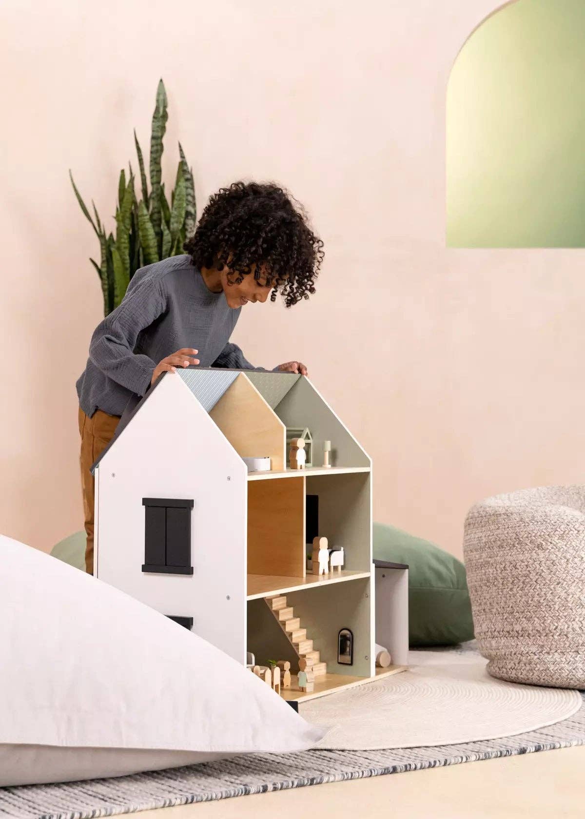 Child playing with a dollhouse in a cozy room with a plant and lamp.