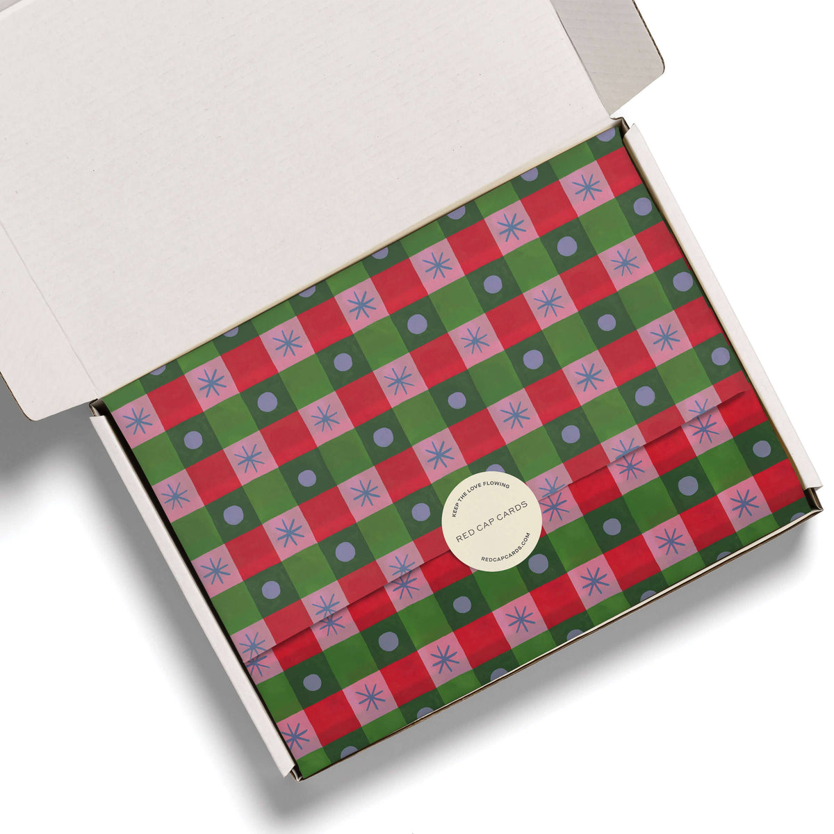An open white cardboard box containing a flat envelope-style package wrapped in a red, green, and lavender plaid holiday pattern with a round white sticker seal.