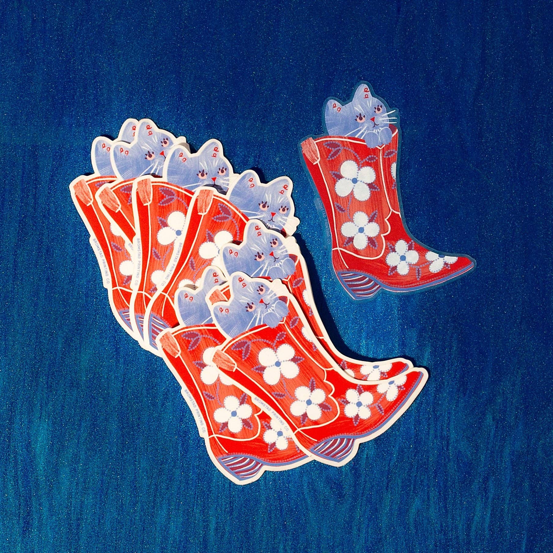 A set of red cowboy boots with white floral patterns and small blue cats peeking out from inside, arranged on a textured blue background.