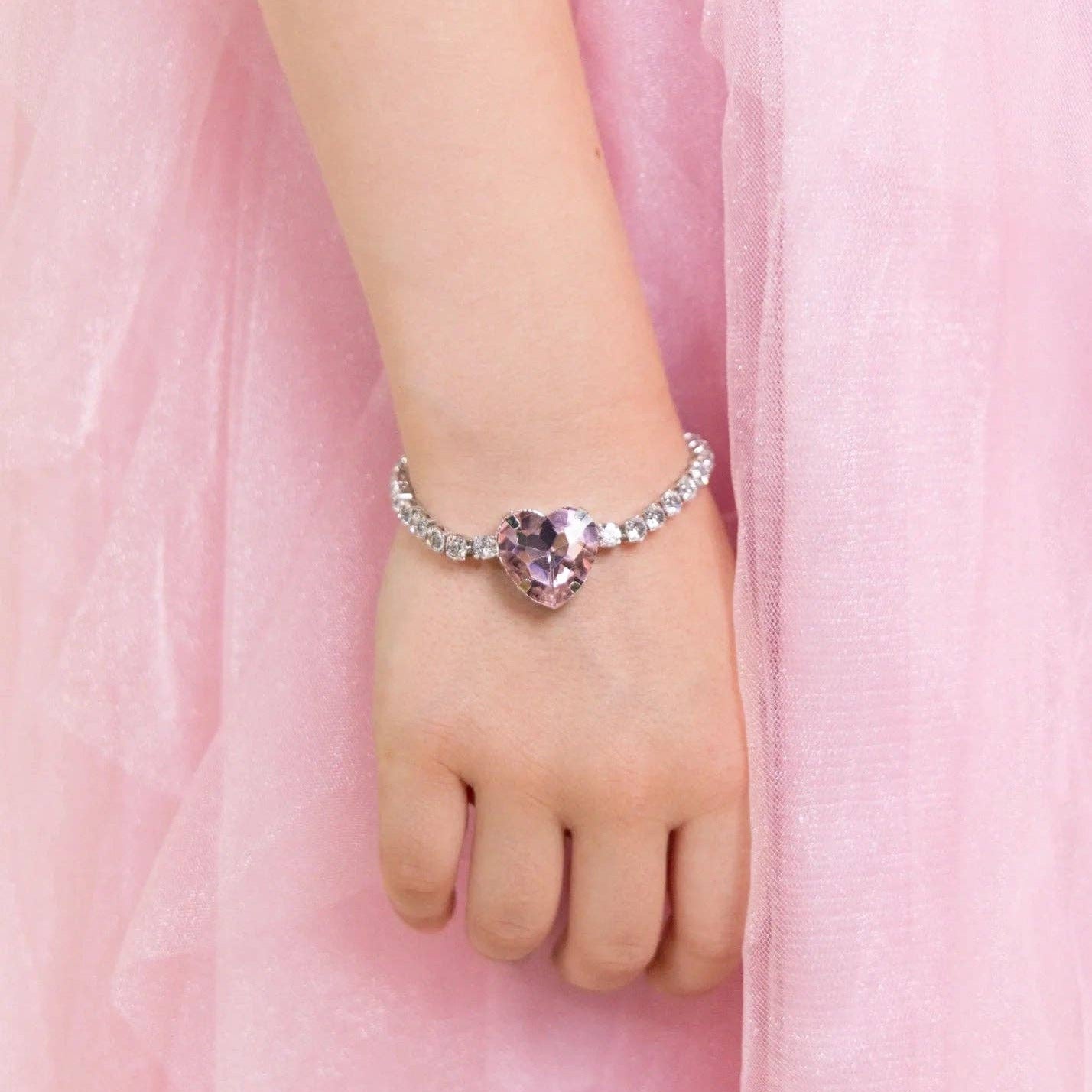 A close-up of a child's hand and wrist wearing a delicate silver-toned bracelet with a heart-shaped purple gemstone, set against a soft pink tulle dress background.