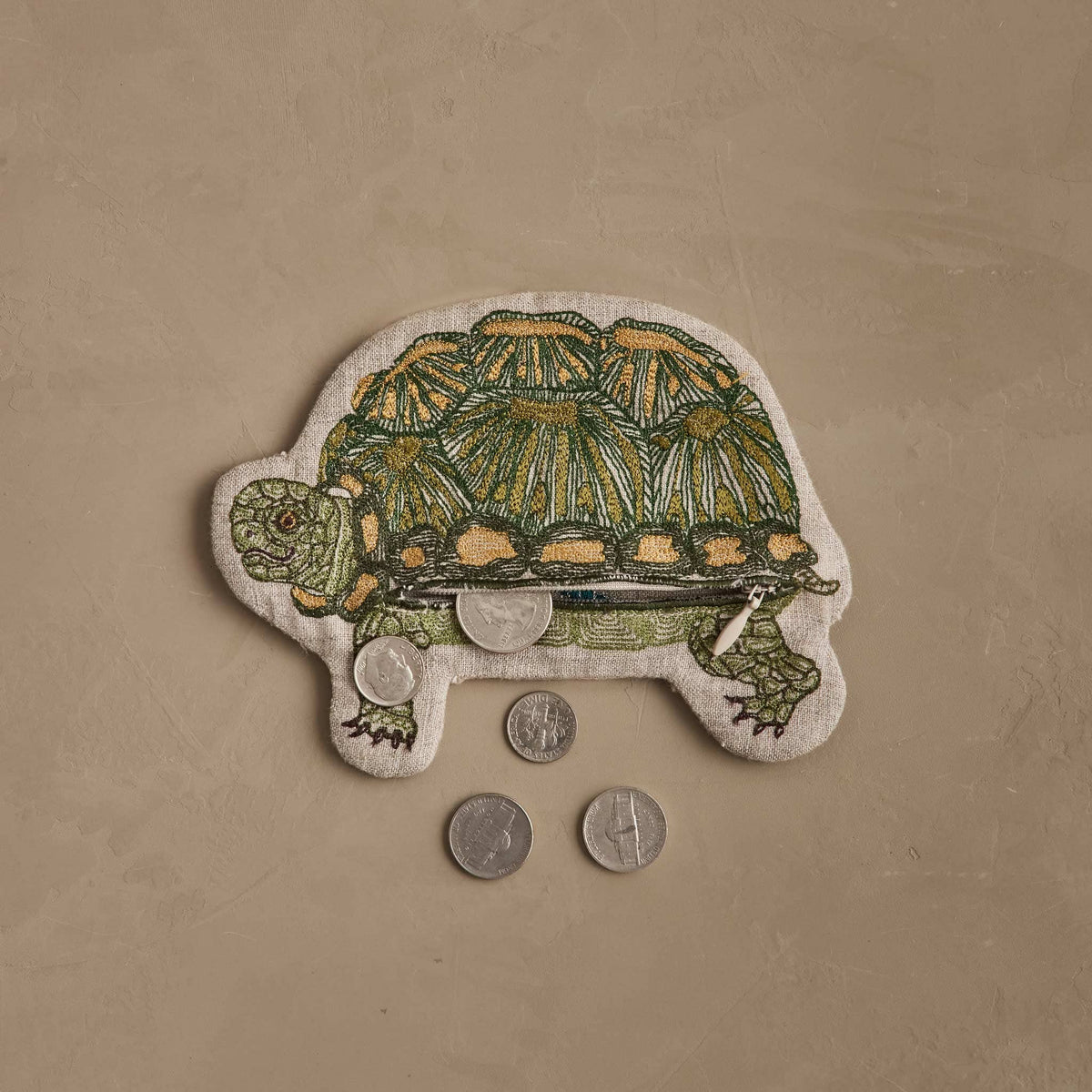 An embroidered turtle patch with a green and yellow shell and head on a white fabric base, displayed on a textured beige background with three coins below for scale.