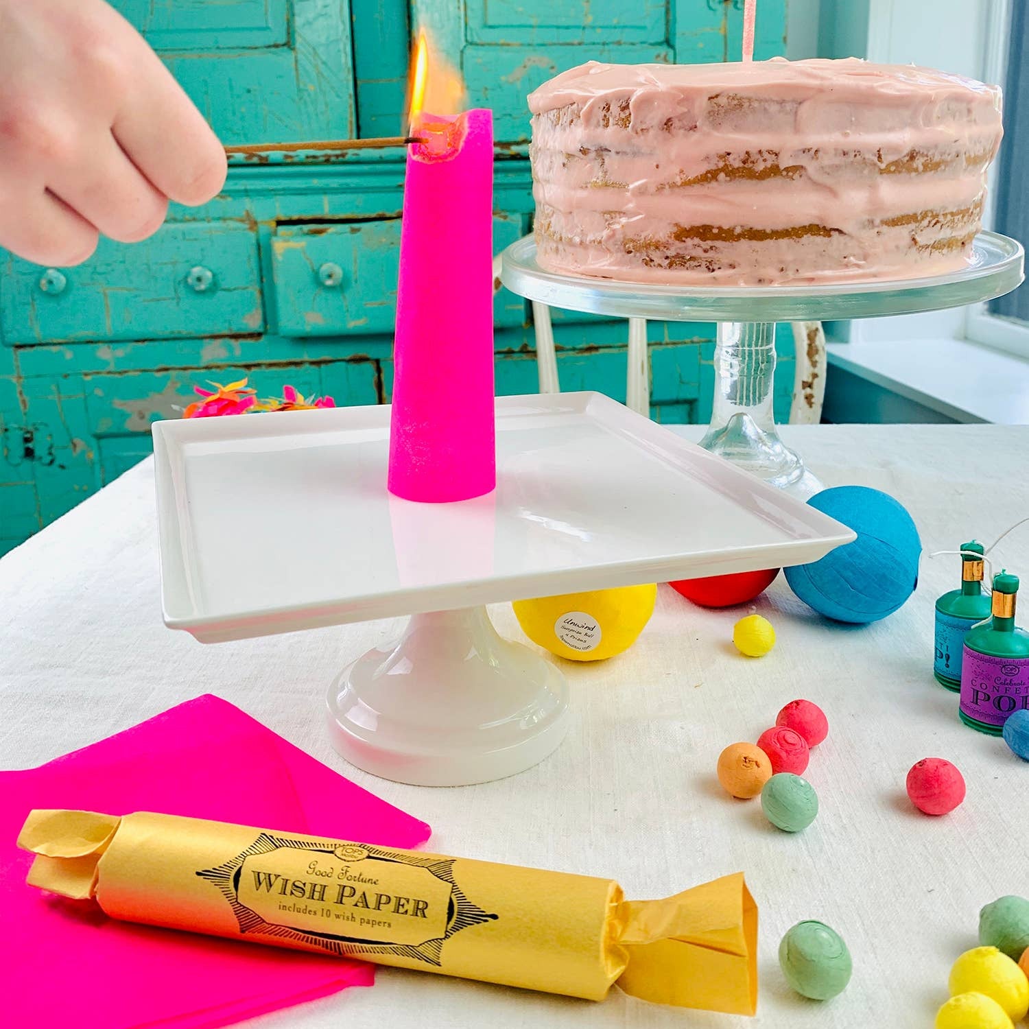 Pink cake with a lit piece of paper on a white plate, surrounded by colorful balls and a roll of wish paper against a turquoise wooden background.