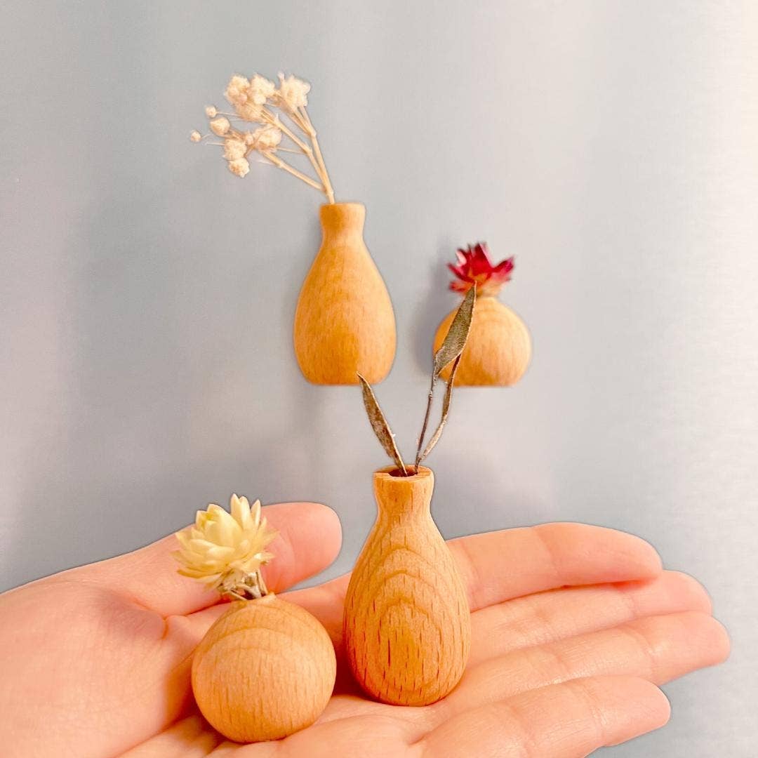 Three small wooden vase-shaped ornaments or charms in a light natural wood finish, displayed on an open hand with dried flowers inserted into two of them, against a soft light gray background.