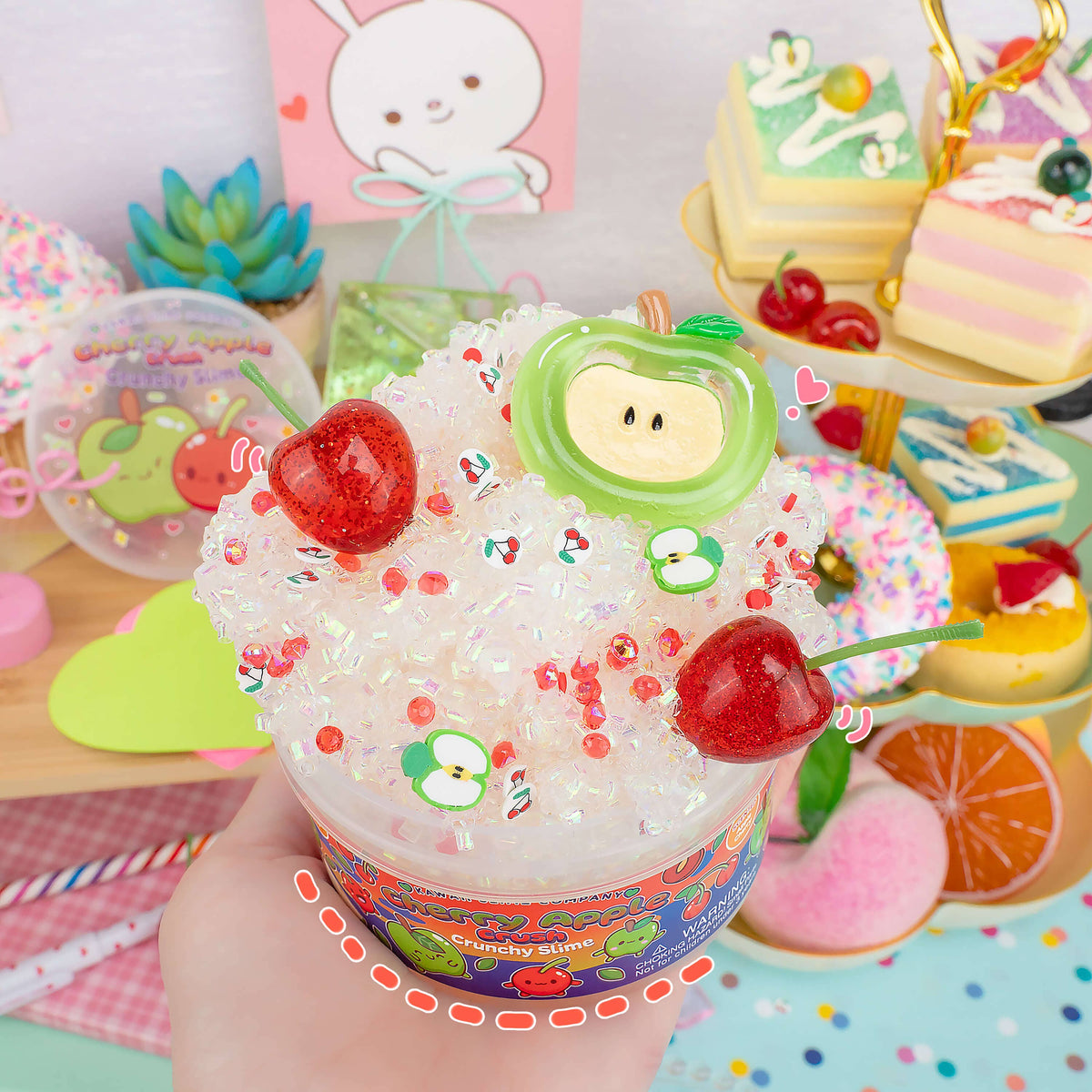 A hand holds a cup of decorative crushed ice or snow-like candy mix topped with two red cherries, green apple-shaped candies, and small red sprinkles. The background is a colorful tabletop with toy desserts, a small succulent, and pastel props.