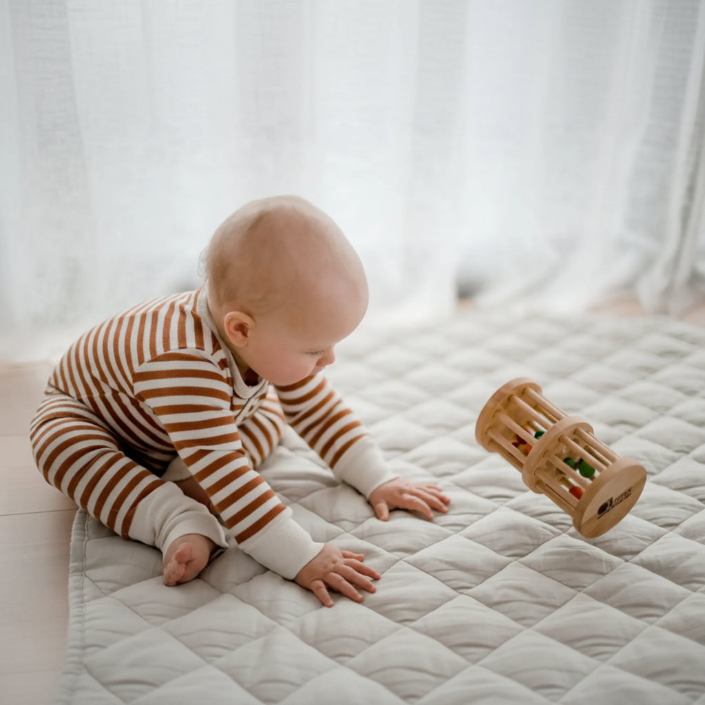 A baby wearing a brown and white striped long-sleeve bodysuit crawls on a quilted cream play mat, looking at a wooden toy.