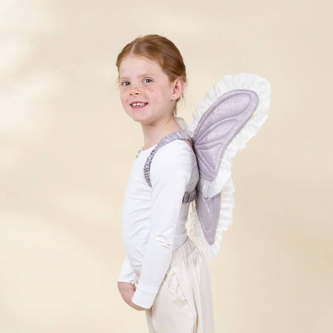 A young girl with red hair stands in profile wearing a white angel costume with white wings that have a light purple border and ruffled trim, set against a warm beige studio background.