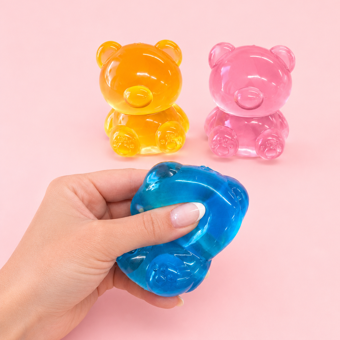 A hand holds a translucent blue bear-shaped stress toy in the foreground, with two more bear-shaped stress toys in yellow and pink blurred in the background against a solid pink backdrop.