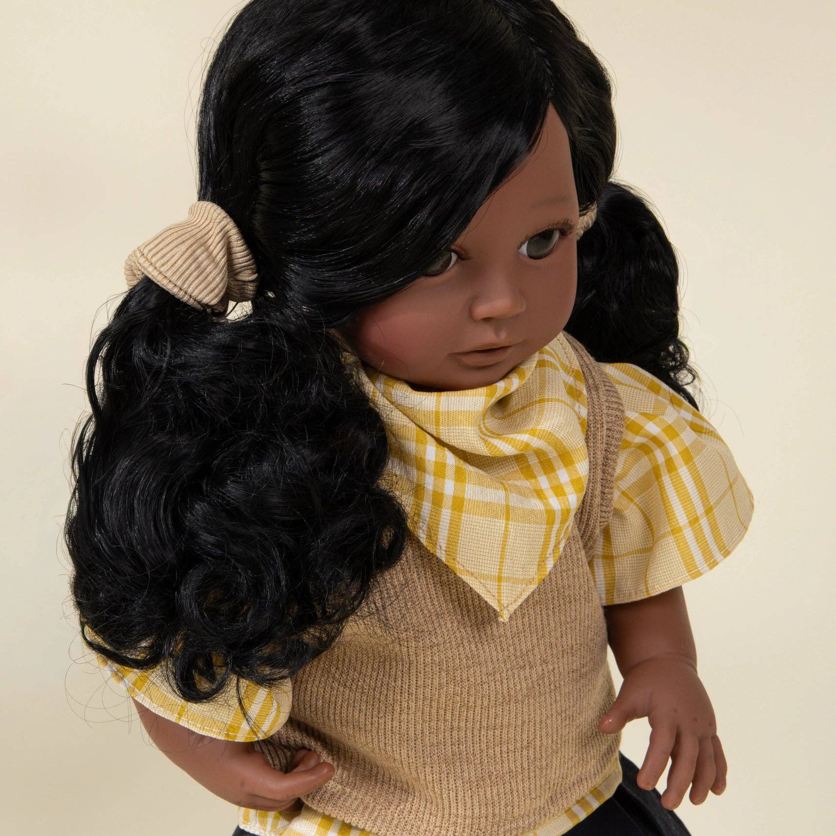 Doll with black hair styled in pigtails, wearing a yellow checkered shirt and brown vest against a beige background.