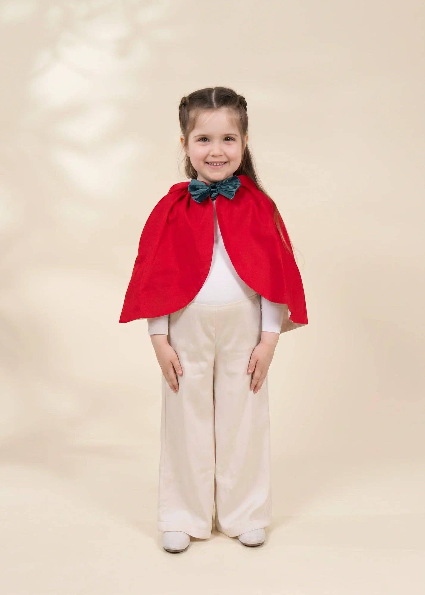 A young girl stands against a warm beige studio backdrop wearing a bright red cape with a dark teal bow at the neckline, a white long-sleeve top, and cream wide-leg pants.